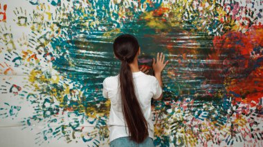 Genç ve güzel bir kızın arka görünüşü renkli duvarı boyamak için elini kaldırır. Çekici bir lise öğrencisi renk paletini tutarken duvarı el iziyle boyuyor. Yaratıcı aktivite. Öğretim.