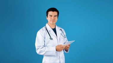 Laboratuvar önlüğü ve steteskobu olan ve mavi arka planda duran profesyonel bir doktor. Kameraya poz verirken tıbbi verilere bakıp kontrol eden yetenekli bir sağlık çalışanı. Yaratıcılık.