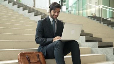 Kendine güveni tam bir iş adamı başarılı iş fikrini müzik dinlerken kutluyor. Happy yönetici projesi kulaklık ve takım elbise giyerken dizüstü bilgisayarla çalışıyor. Yatırımcı merdivenlerde oturuyor. Sevinçli..