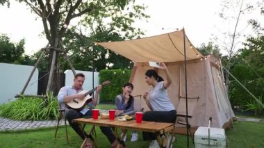 Baba gitar çalarken anne ve kızı bahçede kamp yapıyor. Ebeveynler, genç nesil çapraz kuşak farklılıklarıyla zaman geçirmek için açık hava kamp aktivitelerini kullanıyorlar. Uyumsuzluk.