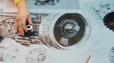 Bu resim, mühendislik ve tasarım sürecinin detaylarını vurgulayarak, bir atölyede teknik çizimlerin yanı sıra karmaşık mekanik parçalar üzerinde çalışan elleri gösteriyor. SACTR
