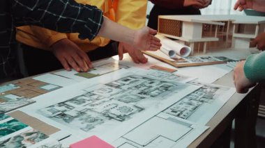 Bir grup mimar bir tasarım oturumuna katılıyor, bina planları, çizimler ve ölçekli modeller üzerinde ellerini gösteriyor, işbirliği ve yaratıcılığı vurguluyorlar. SACTR