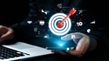 Bir iş adamı akıllı bir telefonla iletişim kurar, dijital ikonlar aracılığıyla hedefleri görselleştirir ve dinamik bir iş ortamında strateji, başarı ve teknolojiyi temsil eder. Dörtgen