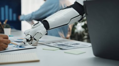 Robotik bir kol modern bir ofiste veri analizi ve çizelge üzerinde çalışan bir profesyonele yardım eder. Bu resim çalışma alanındaki teknoloji ve yeniliğin karışımını vurgular. Noogenesis