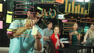 Professional businessman putting sticky notes on glass wall while explain start up project. Skilled project manager pointing at business plan while sharing marketing idea to investor. Tracery