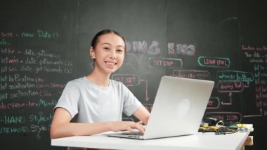 Akademik liseli kız BTMM teknoloji sınıfında kameraya bakarken dizüstü bilgisayar kullanarak kod yazıp çalışıyor. Öğrenci gülümsemesi masaya yerleştirilmiş elektronik aletlerle kameraya. Öğretim