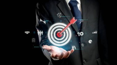 Kendine güvenen bir işadamı dijital bir hedefe kırmızı bir okun merkeze isabet ettiğini gösterir, başarıyı simgeler ve siyah bir arkaplana karşı kurumsal stratejiye odaklanır. Dörtgen