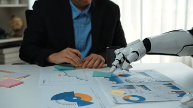 Bir insan ve robotik kol modern ofis ortamında iş birliği yapar, finansal verileri analiz eder ve ticari büyüme ve yenilik stratejilerini tartışır. Noogenesis