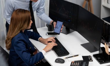 Gülümseyen Asyalı işadamı telefondaki dinamik borsa verilerini bilgisayarda kar değeri değerinin analizi için arayan kadın ortağa gösteriyor. Ofisteki ekranlarla karşılaştırınca. İnfobahn.