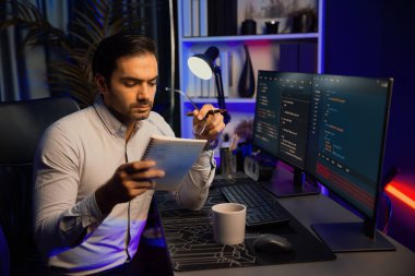 Akıllı bilişim geliştiricisi bilgisayar ekranında yazılım geliştirme kodlaması üzerinde çalışıyor ve en yeni uygulama projesinin notlarını okuyor, program güncellemesi düşünüyor neon ışıklı siber ofiste online web sitesi verileri. Tahmin et..