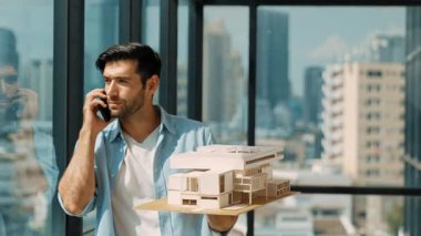 Gülümseyen Kafkasyalı mühendis ev modelini sağa sola çeviriyor ve iş arkadaşına telefon ederken hata noktasını kontrol ediyor. Profesyonel yönetici teftiş, kontrol, mimari modele bakmak. İzleme