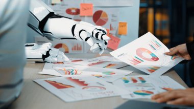 Robotik bir el, modern ofis ortamında bir insanla iş birliği yaparken iş verilerini inceliyor. Bu görüntü analizlerdeki teknoloji entegrasyonunu vurgular. Noogenesis