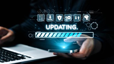 Bilgisayarın yanında, güncelleme ekranı olan akıllı telefonu tutan bir elin yakın görüntüsü, sürekli teknolojik gelişmeyi ve yazılım yönetimini gösteriyor. Dörtgen