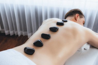 Lüks tatil köyündeki kaplıcada sıcak taş masajı. Gün ışığı dinginliği ortamı, mutlu erkek müşteri bazalt taşı masajının keyfini çıkarıyor. Rahatlatıcı bir sıcaklıkla vücudun üzerinde süzülüyor. Sessiz.