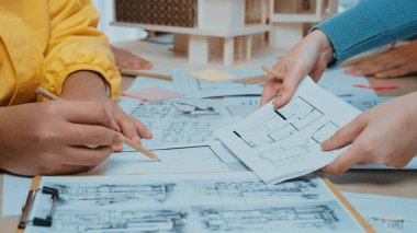 Dinamik işbirlikçi bir atmosferde mimari tasarımlar, çizimler ve planlar üzerinde çalışan bireyler sergileniyor. Nişanlı eller, takım çalışmasının ve yaratıcılığın altını çizer. SACTR