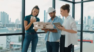 Proje yöneticisi ev modelini elinde tutarken, mimar mühendis takımı da ev tasarımı veya bina inşaatı hakkında fikir alışverişinde bulunuyor. Şehrin penceresinin yanında iç mimar duruyor. İzleme