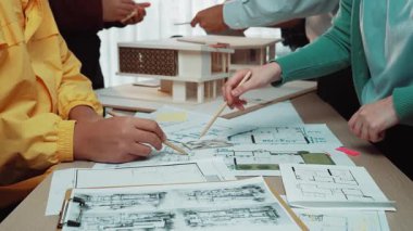 Tasarımcılardan oluşan dinamik bir ekip, mimari modeller ve planlar üzerinde çalışarak modern bir çalışma alanında işbirliği yapıyor. Sahnede profesyoneller arasında yaratıcılık ve takım çalışması sergileniyor. SACTR