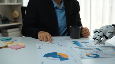 Bir insan ve bir robot el, teknolojiyle dolu modern bir çalışma alanında veri grafikleri ve yenilikçi stratejileri tartışan dinamik bir iş toplantısında işbirliği yaparlar.