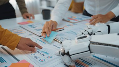 Çağdaş bir ofis ortamında bir insan eli ve bir robot kol işbirliği yapar. Sahne, yeniliği sembolize eden grafikler ve veri analizi ile etkileşimi vurgular. Noogenesis