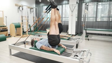 Müşteri, spor salonundaki yenilikçi bir makinede kayışla ters bacak kaldırma hareketi yapıyor. Çekirdek bağlantısını, dengesini ve esnekliğini geliştirmek için ters takla atan iki mutlu antrenör. Alışkanlık.