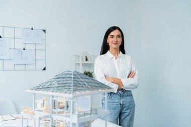 Genç, güzel, profesyonel mimar mühendisinin yakın portresi kollarını kavuşturuyor ve ev modeliyle güvenli bir şekilde beyaz modern ofiste taslak ile poz veriyor. Yaratıcı yaşam ve tasarım. Tertemiz..