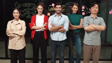 Profesyonel iş ekibi cam duvarda yan yana dururken kollarını kavuşturuyor. Bir grup iş adamı kollarını kavuşturmuş kameraya gülümserken gece ofisinde güvenle duruyorlar. İzleme