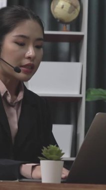 Kulaklık ve mikrofon takan iş kadını masasında çalışıyor ve dizüstü bilgisayar kullanıyor. Uzaktan kumandalı online toplantı için dizüstü bilgisayar kullanan hevesli kadın çalışanlar, ofiste iş videosu konferansı.