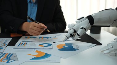 Bir insan ve robot el modern bir iş toplantısında işbirliği yapıyor. Tablodaki tablolar ve grafikler veri analizi ve stratejik planlamayı gösteriyor. Noogenesis