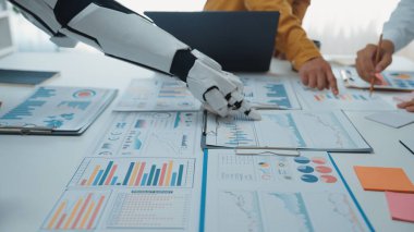 Robotik bir el, modern bir çalışma alanında çeşitli iş haritalarını ve finansal verileri analiz ederek teknoloji ve insan çabaları arasındaki işbirliğini gösterir.