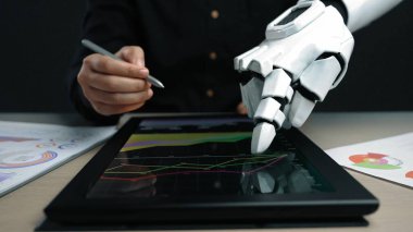 Bir robot el tablet ekrandaki dijital verilerle etkileşime girerken, bir insan eli de teknoloji ve insan çabaları arasındaki işbirliğini temsil eden iş grafikleri üzerine notlar yazıyor.