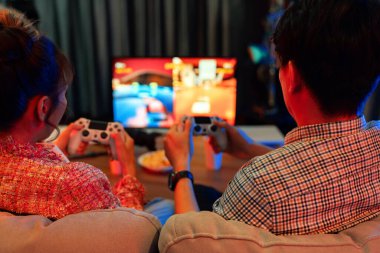 Oyun çubuğuyla oynayan bir çift televizyon ekranında kavga oyunu oynuyor, arka tarafta zorluk derecesi alıyor, modern konforlu evde neon ışıklı oturma odasında kanepeye oturuyor. İnfobahn.