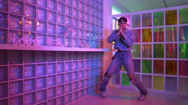 Gece kulübünde kameraya bakarken güneş gözlüklü Asyalı dansçı serbest dans ediyor. Mutlu sokak dansçısı dans ediyor ve renkli parlak neon ışığı ya da yol gösteren ışıkla ayak seslerini gösteriyor. Mutluluk.