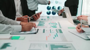 Bir iş toplantısında yer alan çeşitli profesyonellerden oluşan bir grup, takım çalışmasını vurgulayarak, modern bir ofis ortamında kağıt ve cihazlar üzerinde sunulan tablo ve grafikleri analiz ediyor. SACTR