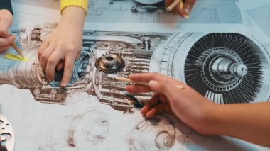 Bir grup katılımcı şahıs, mekanik bileşenlerin karmaşık tasarımları, takım çalışması ve yenilikleri üzerinde çalışmak için araçlar ve planlar kullanarak bir atölyede işbirliği yapıyor. SACTR