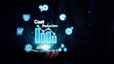 İş dünyasından bir profesyonel, tasarruf ve analizi sembolize eden dijital simgelerle birlikte maliyet düşüşünün grafiksel bir temsilini gösteren bir akıllı telefon tutuyor. Dörtgen