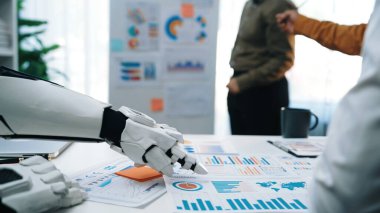 Robot bir kol, modern ofis ortamında gelişmiş teknolojiyi gösteren iş raporlarıyla etkileşime girer. Profesyoneller veri analizi ve içgörü konusunda işbirliği yaparlar. Noogenesis