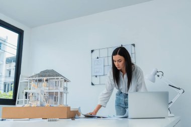 Genç, beyaz, güzel bir mimarın portresi toplantı masasına yerleştirilen ev modeli, plan ve dizüstü bilgisayarın yapısını karşılaştırmaya odaklanıyor. Şah, teftiş, tahmin konsepti. Tertemiz..