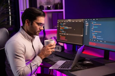 Akıllı IT geliştiricisi bilgisayar ekranında yazılım geliştirme kodlamasında çalışırken kahve fincanı içiyor, neon ışıklandırma siber ofisinde program uygulama güncelleme veritabanı sunuyor. Tahmin et..