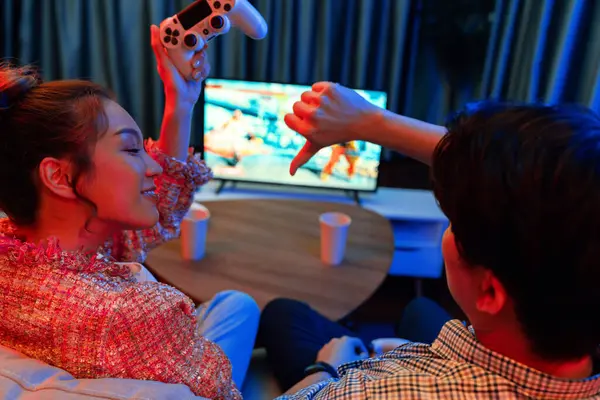 Joystickli birkaç oyuncu dövüş oyunu oynuyor, kadın oyuncu yarışması derecesi arka tarafta TV ekranında modern rahat evinde neon ışıklı oturma odasındaki koltukta oturuyor. İnfobahn.