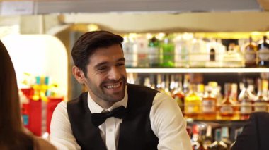 Güzel görünümlü barmen, içki tezgahındaki çiftle samimi bir gülümseme sohbeti yaparken aynı zamanda hoş geldin içkisi ve özel menü listesi öneren çekici profesyonellik. Vinosity.