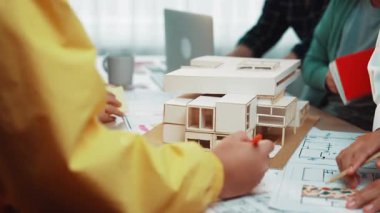 Modern bir ofiste dinamik takım toplantısı, mimari tasarım ve stratejiye odaklanma, bir model, planlar ve işbirliği için masaya saçılmış araçlar. SACTR