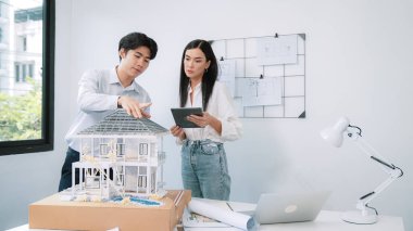 Güzel, genç, beyaz bir proje yöneticisi Asyalı mühendise, mühendislik masasında ev modelini ölçerken tablet kullanarak bina detaylarını anlattı. Tertemiz.