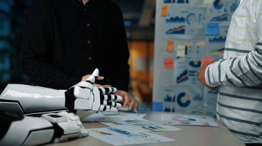 Dinamik bir iş toplantısı. Robot bir elin yanında olduğu, grafikleri ve grafikleri analiz eden, modern bir ofiste teknoloji ve takım çalışmasının karışımını vurgulayan. Noogenesis