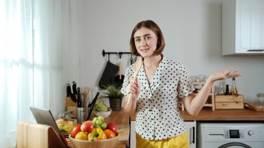 Kafkasyalı anne, modern mutfakta dizüstü bilgisayarda yemek tarifine bakarken salata yapmak için yemek hazırlıyor. Yetenekli ev bekçisi taze sebze kullanarak kahvaltı hazırlıyor. Pedagoji.