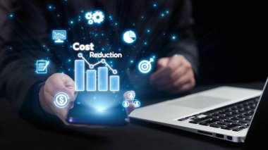 Bir işletme profesyoneli, maliyetlerin azaltılması ile ilgili dijital grafik ve simgeleri gösteren bir akıllı telefon sergileyerek, modern araçları ve finans alanında stratejik planlamayı vurguluyor. Dörtgen