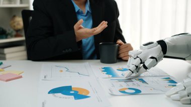 Bir insan ve bir robot ofis masasında etkileşime girer, kahve içerken çizelgeleri ve veri raporlarını birlikte inceler, iş analizlerinde modern işbirliği sergilerler.