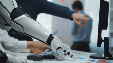 Robot bir kol, modern ofis ortamında bir bilgisayarla etkileşime girer, bu da teknoloji ve insan çabalarının iş yerinde bütünleşmesini sembolize eder. Noogenesis
