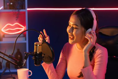 Genç ve güzel Asyalı yayıncının sunucu kanalı, dinleyicilerle sosyal medyada müzik dinliyor, pastel kulaklık takıyor, kapalı alanda neon ışıklı stüdyoda şarkı söylemek için mikrofon kullanıyor. Taktik.