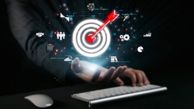 Bir işadamı, karanlık bir arkaplana karşı strateji, teknoloji ve takım çalışmasını temsil eden çeşitli simgelerle çevrili dijital hedef grafiğine uzanıyor. Dörtgen