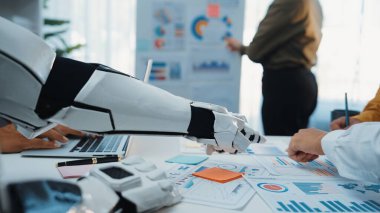 Bir robot kolu, modern ofis ortamında çizelgeleri ve planları analiz eden profesyoneller tarafından çevrelenmiş veri analizlerine odaklanmış bir iş toplantısında yardımcı oluyor.